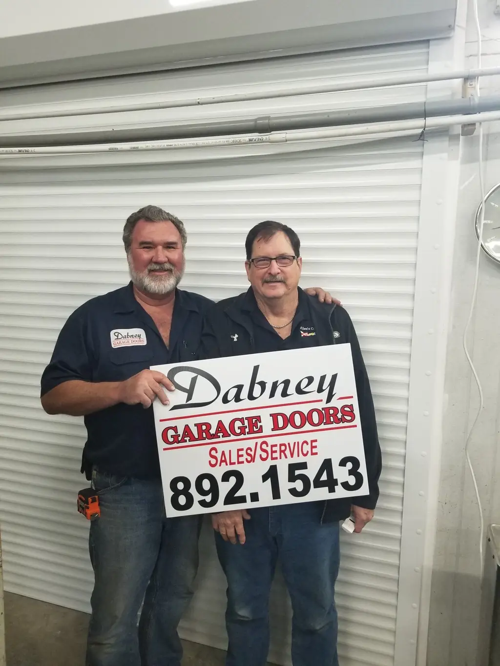 Two Dabney Garage Doors installers in company shirts holding a Dabney sign board in the shop bay